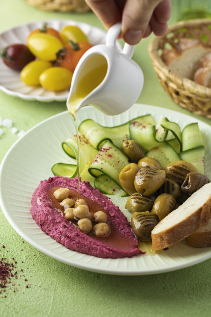 Vegan Mediterranean platter featuring beet hummus, chickpeas, grilled olives, cucumber ribbons, and fresh bread with olive oil drizzle.