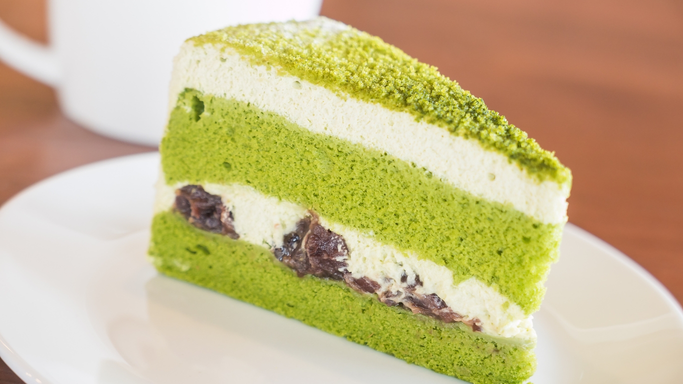 Slice of vibrant vegan matcha cake with green sponge layers, creamy frosting, and sweet red bean filling, served on a white plate.