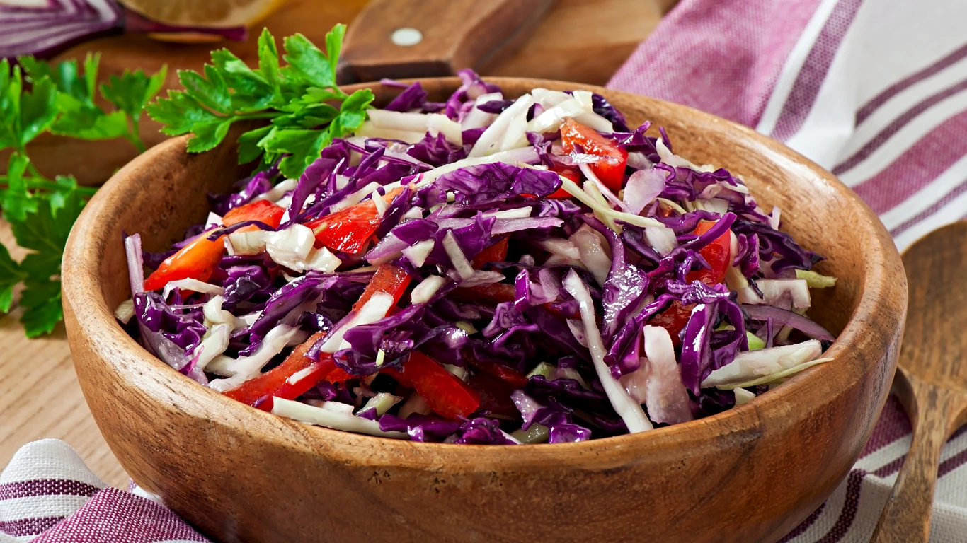 A wooden bowl filled with vibrant vegan coleslaw made of shredded purple cabbage, red bell peppers, and white cabbage, garnished with fresh parsley, set on a rustic table with a striped kitchen towel and wooden spoon in the background.