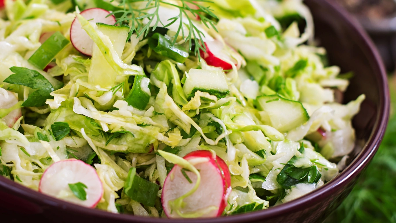 Close-up of a fresh, vibrant vegan coleslaw with shredded cabbage, radishes, cucumbers, and green herbs, all in a dark bowl.