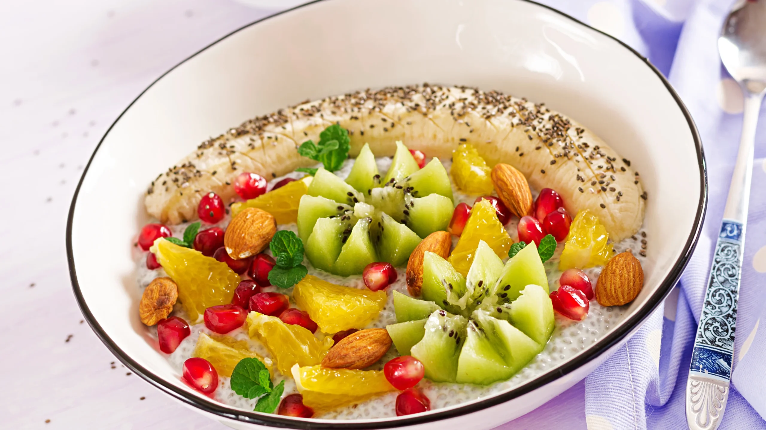 A vibrant vegan breakfast bowl featuring sliced banana topped with chia seeds, fresh kiwi arranged in flower shapes, pomegranate seeds, orange slices, almonds, and garnished with mint leaves, served in a white bowl on a light background with a decorative spoon.