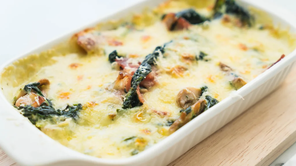 Close-up of a golden-baked vegan casserole with spinach, mushrooms, and a creamy topping in a white ceramic dish.