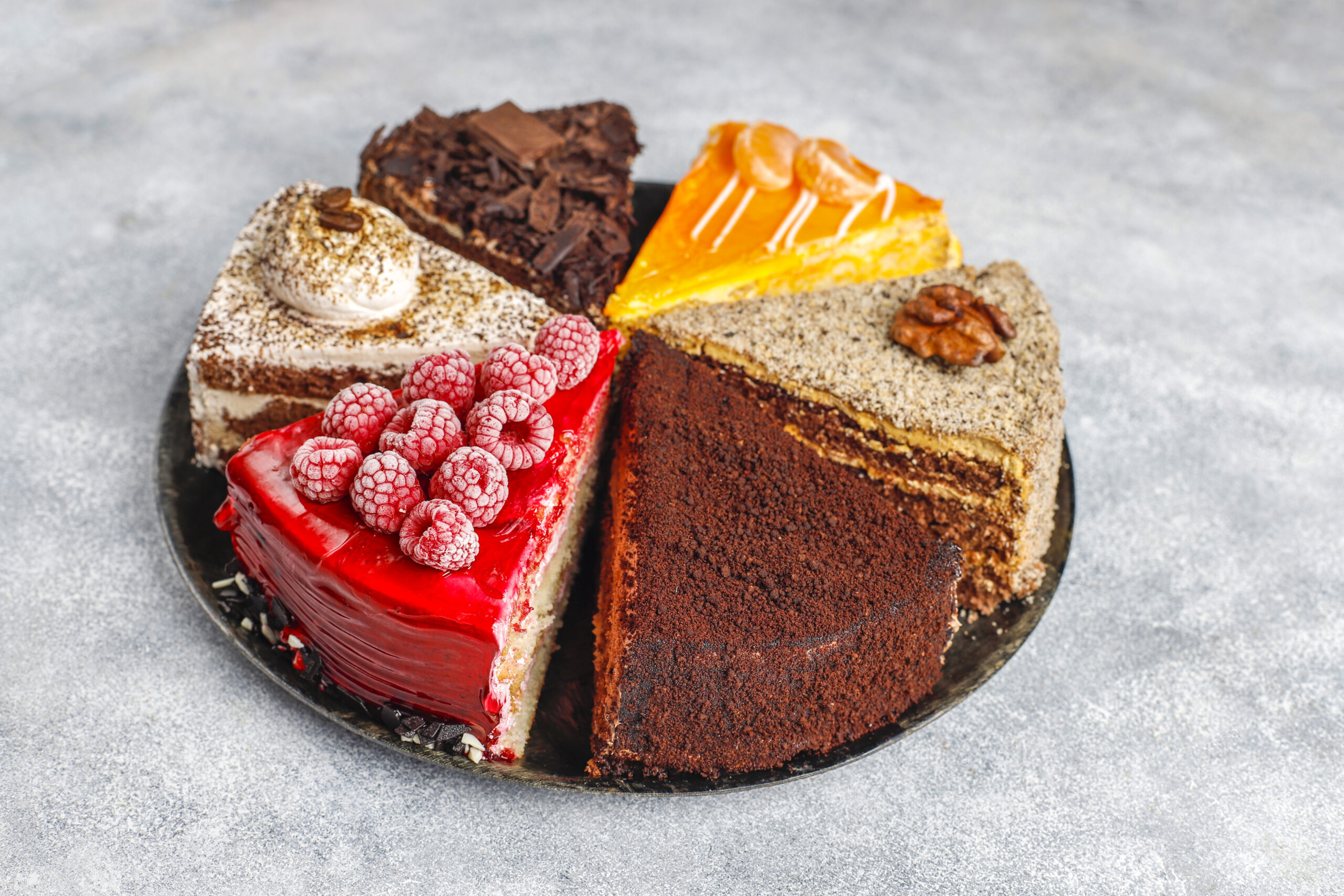 An assortment of colorful cake slices, including chocolate, raspberry, orange, and nut-topped options, arranged on a round plate on a light textured background.