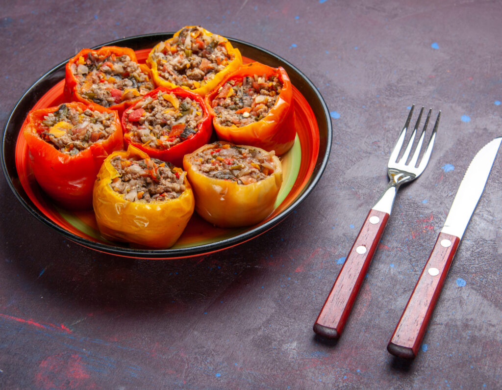 Vegan stuffed bell peppers filled with a savory plant-based mixture, served on a decorative plate with a fork and knife on a textured dark surface.