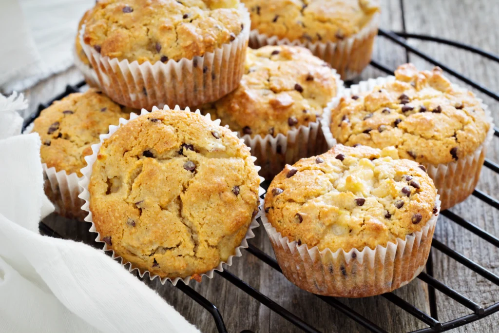 A close-up of freshly baked vegan banana muffins with chocolate chips on a black wire cooling rack. The muffins are golden-brown with a cracked, fluffy texture, sitting on a rustic wooden table with a white napkin in the background