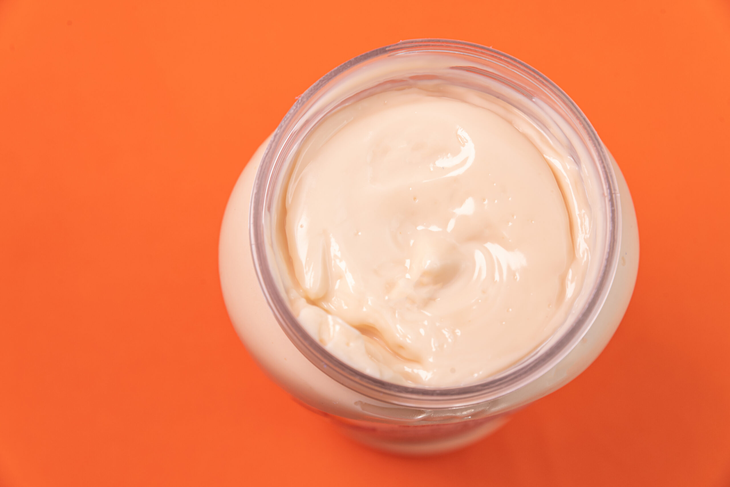 A jar of creamy mayonnaise with a smooth texture, photographed against a vibrant orange background.