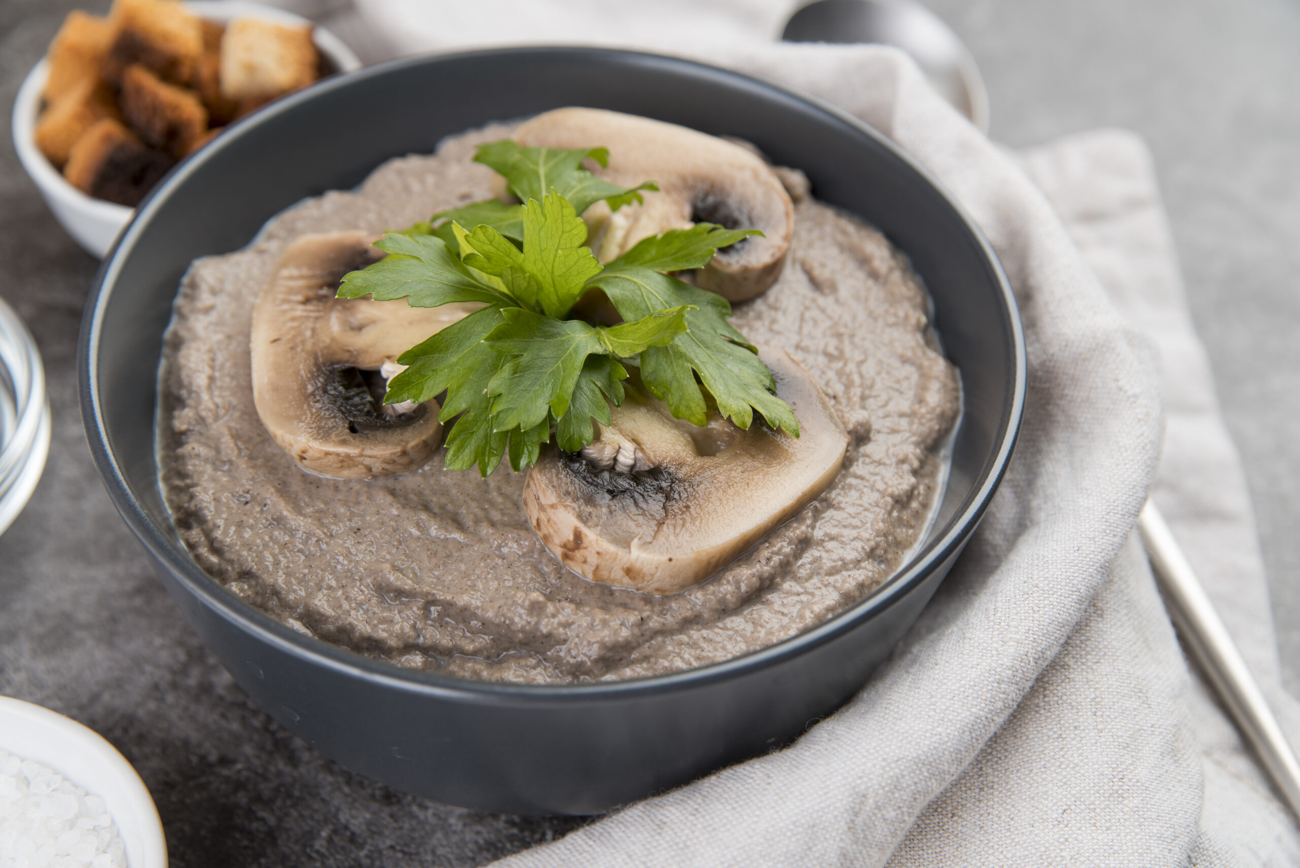 Vegan mushroom cream soup garnished with parsley and fresh mushroom slices, served in a black bowl on a gray napkin.