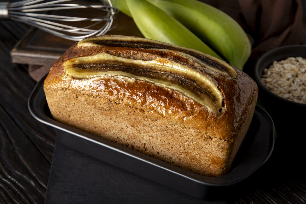 Freshly baked banana bread in a loaf pan, topped with sliced bananas, placed on a dark wooden table with green bananas, oats, and a whisk in the background.