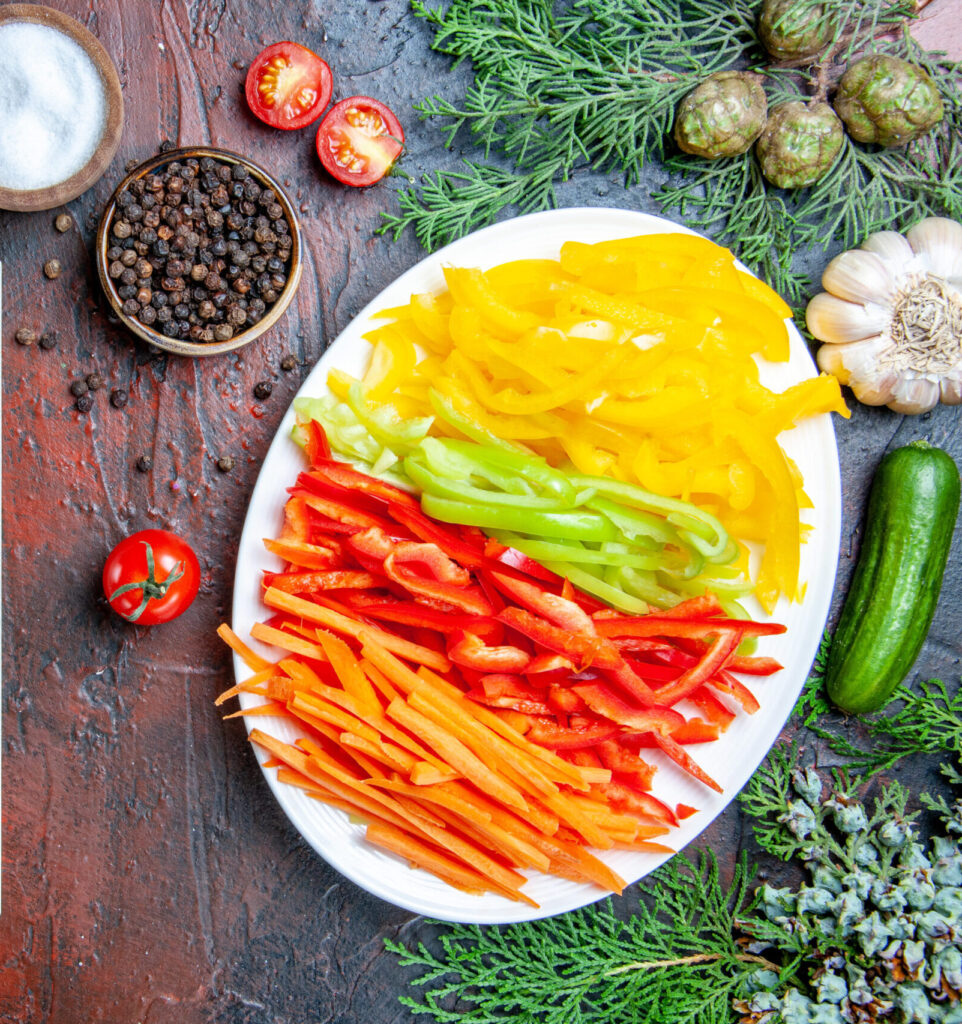 Top view of a raw vegan recipe setup featuring colorful sliced peppers on a white plate, fresh cherry tomatoes, garlic, cucumber, spices, and a notepad with a pencil on a textured dark red table.