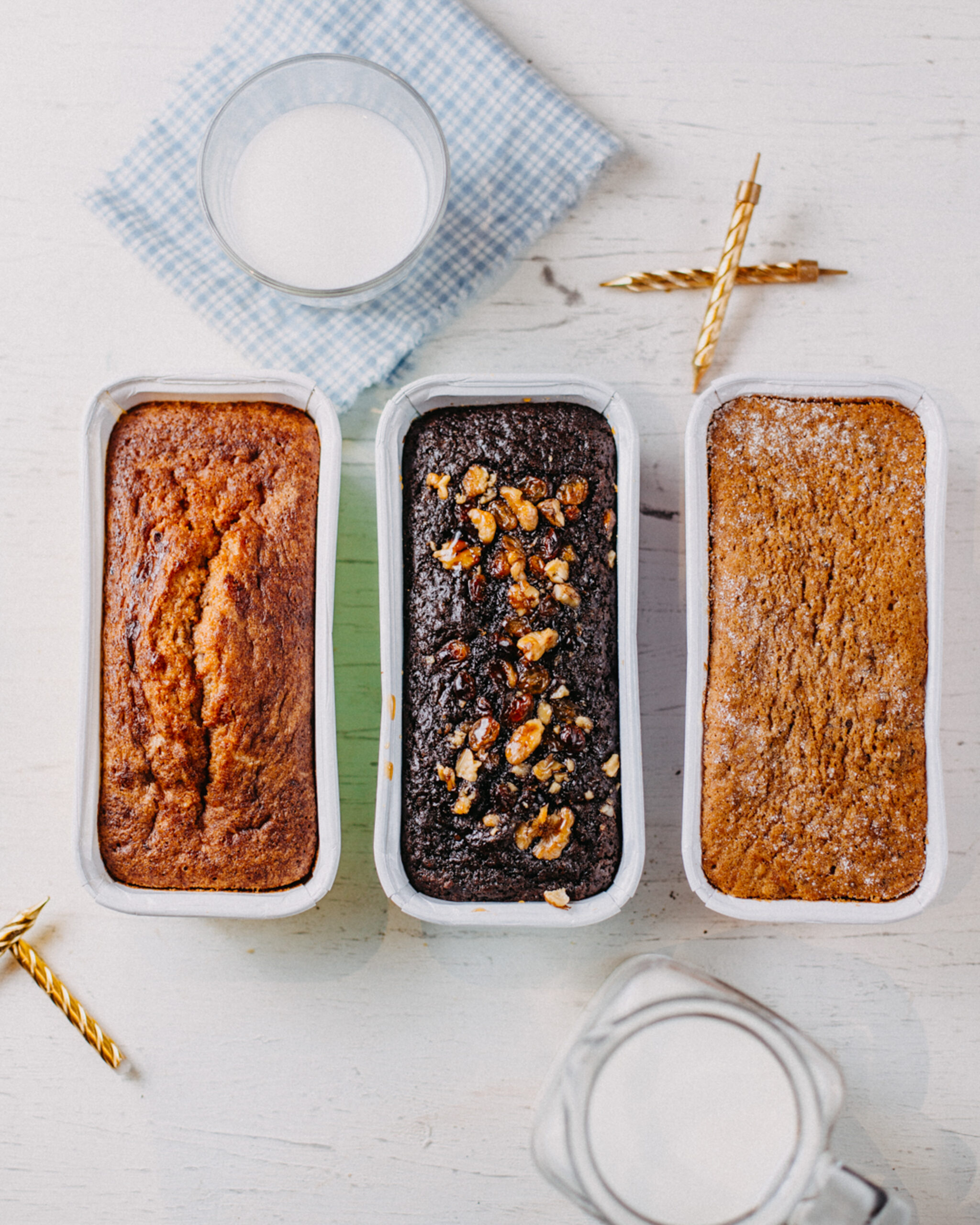 An assortment of three homemade vegan cakes in loaf tins, featuring chocolate and fruit flavors, with a glass of plant-based milk in the background.